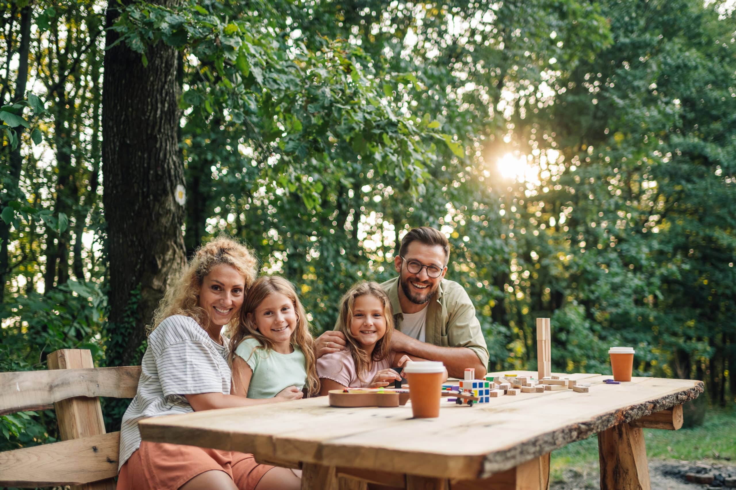 Portrait of a loving family sitting at the table in nature, hugging and smiling at the camera. A cheerful family having a board game day in nature. Parents hugging daughters and looking at the camera.